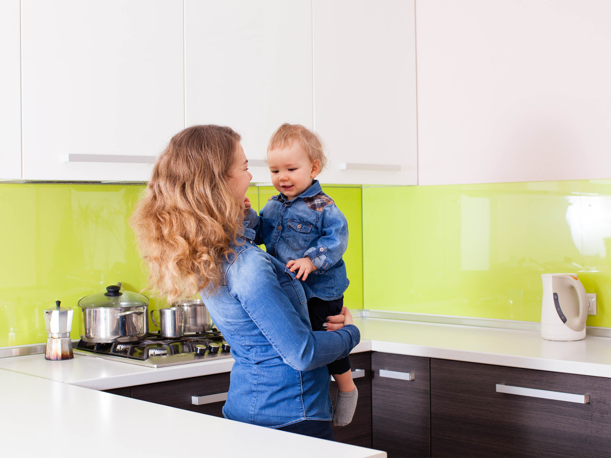 Mutter mit Kind in moderner Küche Eine Frau mit lockigem Haar hält ihr kleines Kind in einer modernen Küche mit grüner Rückwand und weißem Wasserkocher.